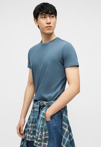 Junger Mann, der ein blaues T-Shirt und Jeans trägt, mit einem grün-blau karierten Hemd um die Taille gebunden, schaut zur Seite.