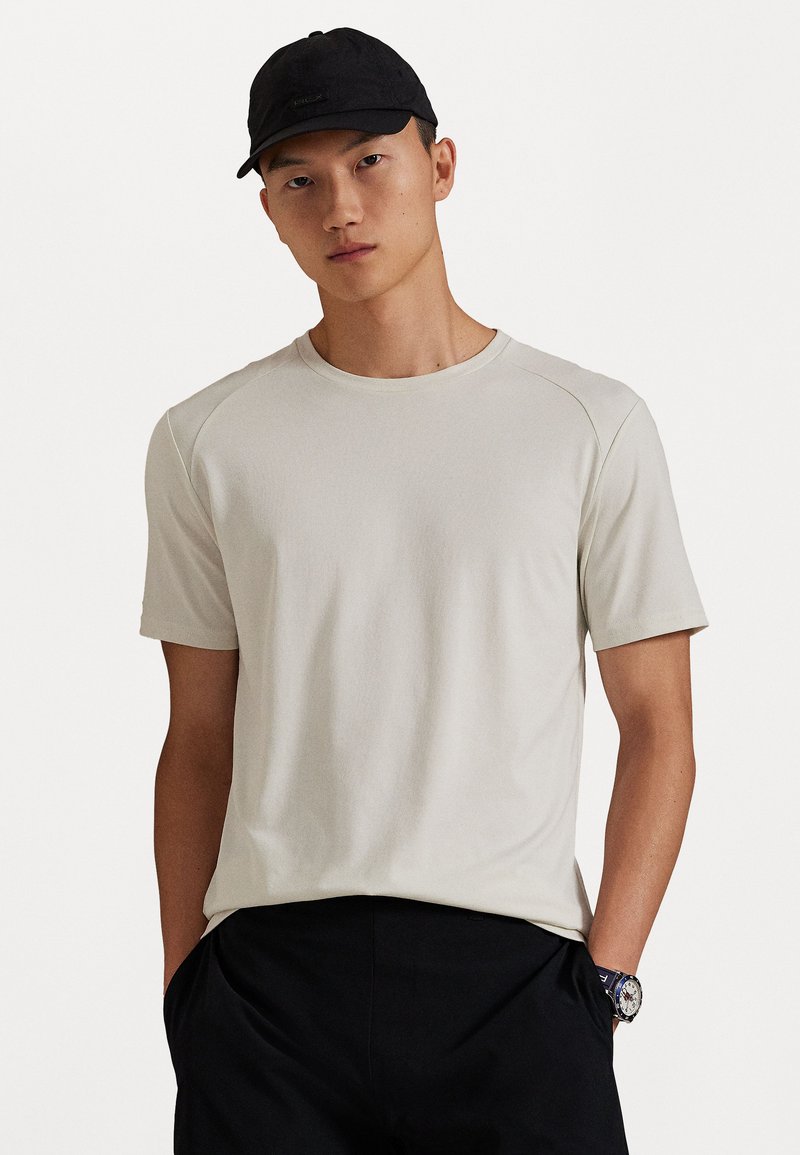 Νεαρός άνδρας ντυμένος με ένα μπεζ πουκάμισο κοντομάνικ�ο, μαύρο καπέλο, μαύρο παντελόνι και ρολόι χειρός, με τα χέρια στις τσέπες του σε απλό φόντο.