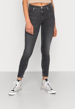 Femme portant un jean skinny noir taille haute et des baskets montantes blanches à taches noires, debout devant un fond uni.