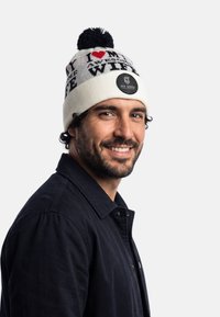 Stickad mössa i krämfärg med svart text som lyder "Jag älskar min fantastiska fru" och en svart pompon; matchad med en mörk jacka.