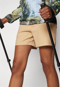 Bermudas beges com um corte reto, design minimalista e um leve brilho. Uma pessoa está a segurar bastões de trekking, vestindo uma camisa com um padrão colorido.
