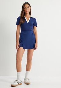 Top de manga corta azul marino con cuello blanco, combinado con una falda plisada azul a juego. La modelo lleva zapatillas blancas y calcetines acanalados.