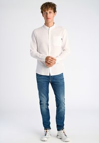 Lichtgekleurde button-up shirt met een staande kraag en een borstzak, gecombineerd met blauwe denim jeans en witte sneakers.