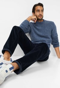 Marineblauwe corduroy broek, lichtblauw gestructureerd brei-trui, witte sneakers met blauwe accenten. Model zit tegen een witte achtergrond.