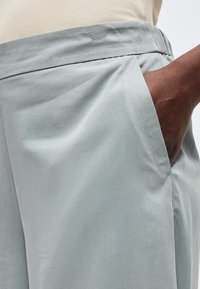 Χέρι με σκούρο δέρμα τοποθετημένο στην τσέπη ανοιχτόχρωμων γκρι παντελονιών που φοριούνται με μπεζ μπλουζάκι, δείχνοντας υφή υφάσματος και λεπτομέρειες ραφής.