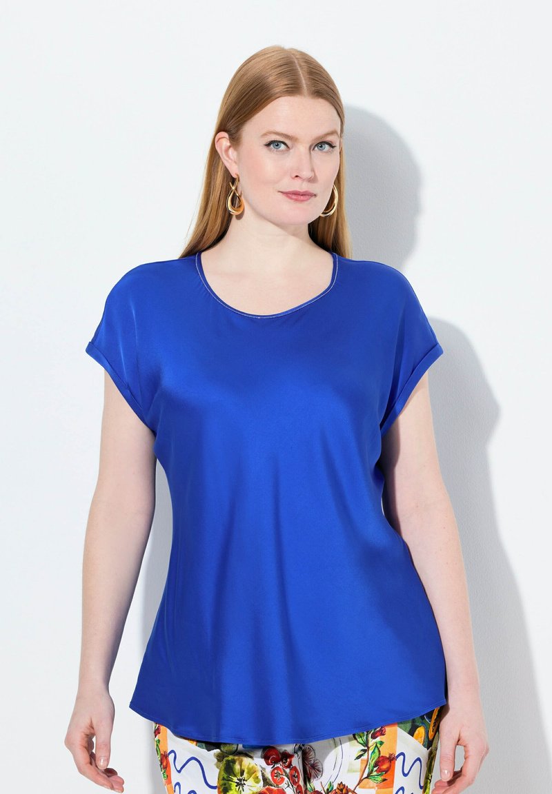 Top blu corto con maniche a sbuffo e scollatura rotonda, caratterizzato da una texture liscia e lucida. Presenta maniche arrotolate e una vestibilità comoda, indossato con pantaloni a fantasia.