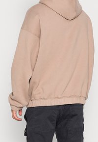 Sudadera con capucha beige de gran tamaño con cordón ajustable, puños y dobladillo acanalados; confeccionada en tela suave y texturizada; vista desde atrás.