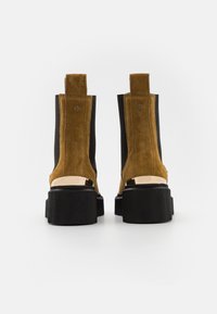 Stivaletti in suede marrone con pannelli elastici neri sui lati, suole in gomma nera spessa e dettagli metallici sul tallone. Vista posteriore mostrata.
