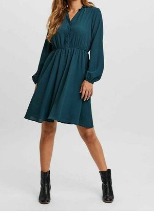 Vrouw die een knie-lange donkerteal jurk met lange mouwen en knopen draagt, samen met zwarte enkellaarsjes, staand met gespreide benen tegen een effen achtergrond.