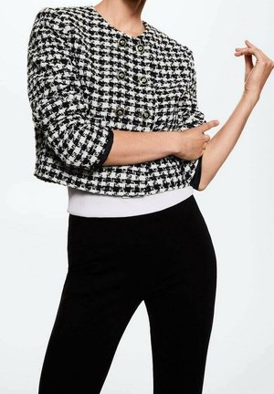 Mujer con chaqueta corta de pata de gallo en blanco y negro con botones y pantalones negros, ajustándose una manga con la mano contraria.