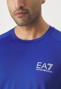 Camisa atlética azul con una tela texturizada, cuello redondo y mangas cortas. Presenta un logo blanco de EA7 Emporio Armani en el pecho.
