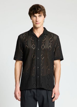 Giovane uomo dalla pelle chiara e capelli castani indossa una camicia nera a maniche corte con motivo geometrico in pizzo e pantaloni scuri su sfondo semplice.