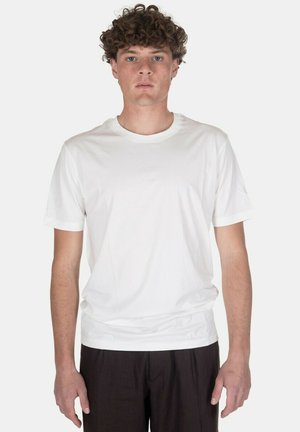 Giovane uomo con capelli ricci che indossa una maglietta bianca semplice a maniche corte e pantaloni scuri, in piedi davanti a uno sfondo grigio chiaro.