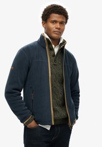Chaqueta de forro polar azul marino con cremallera dorada, cuello alto y bolsillos frontales. Debajo hay un suéter verde texturizado con cuello abotonado.