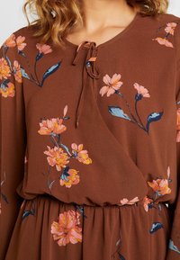 Brun blommig klänning med rosa och blå blommönster, rund halsringning och en knytning fram. Mjuk tyg med uppdragen midja.