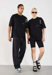 Deux modèles portent des t-shirts noirs en grande taille, associés à des jeans foncés et des shorts. Les accessoires notables incluent des lunettes de soleil et des chaussettes superposées avec des sandales.
