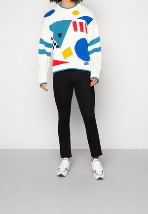 Osoba stojící před čistým pozadím, oblečená v bílém svetru s barevnými geometrickými tvary, černých kalhotách a bílých teniskách.