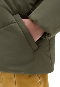 Chaqueta acolchada de color verde oliva con un bolsillo lateral. Presenta un exterior suave y puños elásticos, combinada con unos pantalones de tela amarilla.