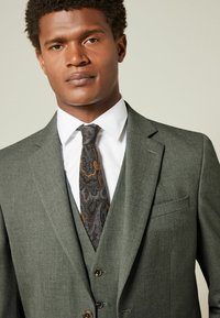 Veste de costume grise avec des revers crantés, en tissu texturé, et poches latérales, portée sur une chemise blanche et une cravate sombre à motifs. Gilet à trois boutons en dessous.