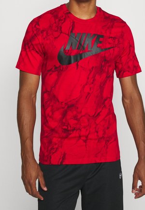 Mężczyzna w czerwonej koszulce Nike z wzorem marmuru i dużym czarnym logo, połączonej z czarnymi spodeńkami Nike.