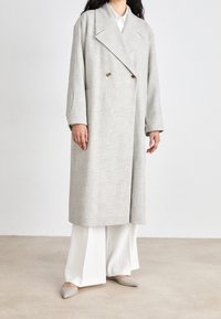 Lichtgrijze oversized jas met een dubbelzijdig ontwerp, grote revers en een subtiel ruitpatroon, gecombineerd met witte wijde broek en platte schoenen.