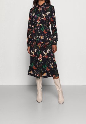 Frau trägt ein langärmliges, vorne geknöpftes schwarzes Kleid mit bunten Blumenmustern und cremefarbenen knielangen Stiefeln, steht vor einem einfarbigen Hintergrund.