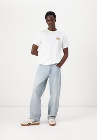 Hvit bomulls t-skjorte med et lite fargerikt grafisk motiv på brystet, kombinert med lyseblå, løstsittende jeans og kremfargede sneakers.