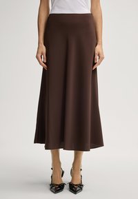 Jupe brune, longue jusqu'au sol, en tissu lisse avec une taille cintrée et un bas évasé. Portée avec des Talons noirs à bout pointu.