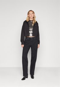 Haut noir à capuche coupé avec une fermeture éclair, top argent métallisé en dessous, jean noir à jambe droite et bottines à talons noires. Design simple.