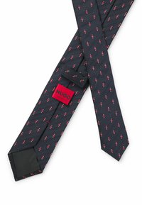 Cravate bleu marine avec un motif en chevrons, ornée de petits motifs rouges et blancs. Comprend une étiquette rouge avec "HUGO" et "FABRIQUÉ EN ITALIE."