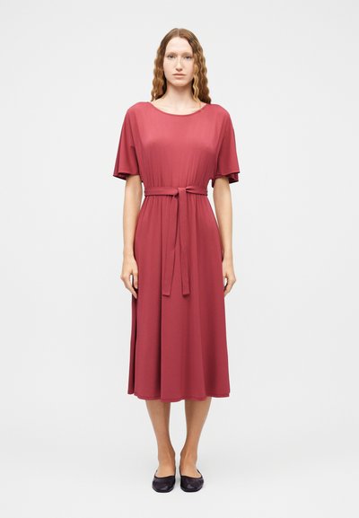 Femme aux cheveux longs et ondulés portant une robe rose poussiéreuse jusqu'aux genoux, à manches courtes et ceinture à la taille, associée à des ballerines noires, debout face à l'avant.