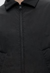 Μαύρο μπουφάν με καπιτονέ, φερμουάρ μπροστά, φαρδύ γιακά και πλαϊνές τσέπες.