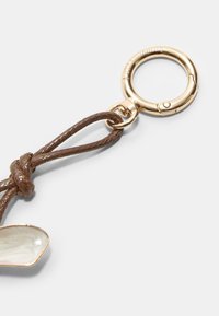 Corda intrecciata marrone attaccata a un anello in metallo tono oro, con un piccolo pezzo decorativo ovale di colore crema con una superficie liscia.