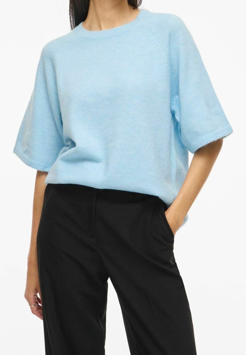 Femme portant un pull bleu clair à manches courtes et un pantalon noir, une main dans la poche, sur un fond blanc uni.
