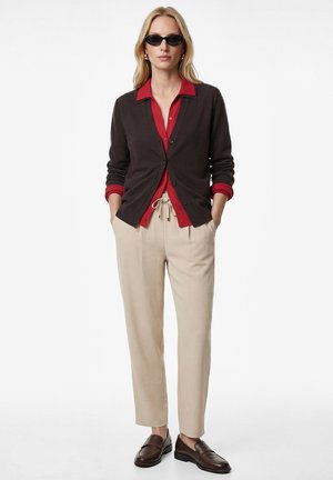 Femme portant un cardigan noir sur une chemise rouge à boutons, un pantalon beige à cordon, des mocassins marron foncé et des lunettes de soleil noires, debout les mains dans les poches.