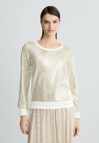 Felpa con paillettes color oro chiaro, maniche lunghe e scollatura rotonda. Presenta polsini e orlo color crema, una texture morbida e una vestibilità rilassata.