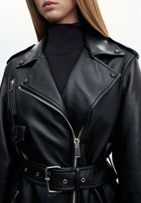 Femme portant une veste moto en cuir noir avec une fermeture éclair et des boutons argentés, ceinturée à la taille, par-dessus un haut à col roulé noir.