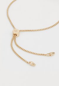 Bracciale regolabile in tono dorato con link di catena fine, un fermo cilindrico con iniziali incise e estremità appuntite per una chiusura sicura.