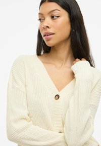 Femme aux longs cheveux bruns portant un cardigan tricoté crème à gros boutons, regardant vers la gauche sur un fond blanc.