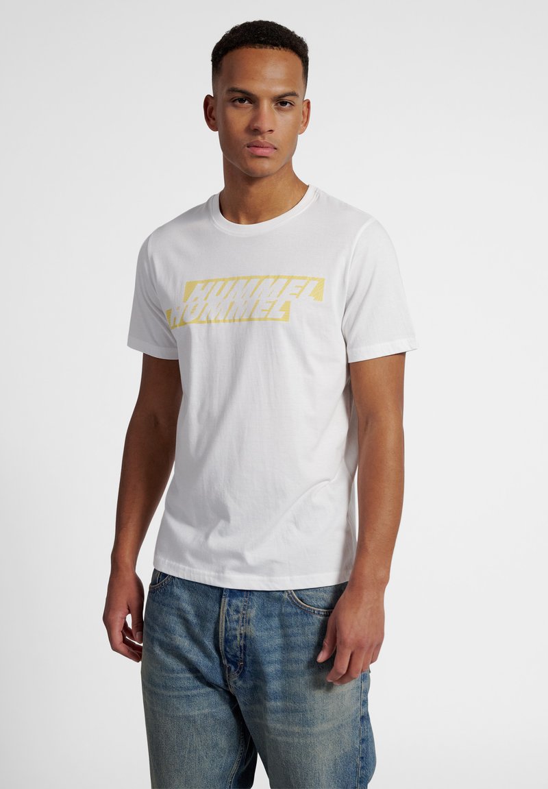 Ung mand iført en hvid t-shirt med gul "HUMMEL"-logo og blå jeans, stående mod en ensfarvet hvid baggrund.