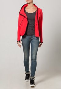 Red zip-up jacket with a hood, black accents, and a diagonal front design, paired with a gray top and fitted blue jeans, worn with black sneakers.