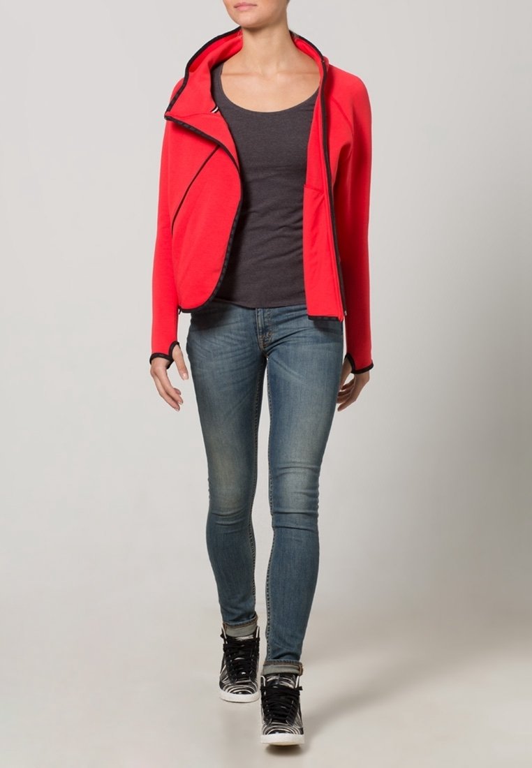 Red zip-up jacket with a hood, black accents, and a diagonal front design, paired with a gray top and fitted blue jeans, worn with black sneakers.