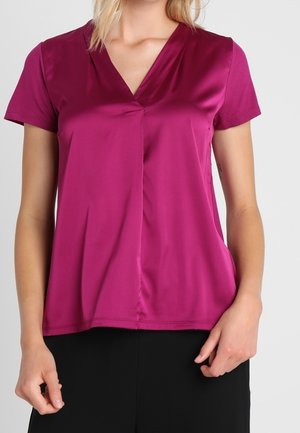 Kobieta ubrana w fioletową bluzkę z krótkim rękawem, dekoltem w kształcie litery V i plisowanym przodem, w połączeniu z czarnymi spodniami na jednolitym tle.