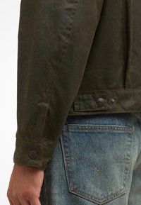 Λαδί κεντημένο μπουφάν με κουμπωτά μανίκια και ορατές ραφές, συνδυασμένο με μπλε τζιν παντελόνι που διαθέτει αντίθετες λεπτομέρειες στις τσέπες.