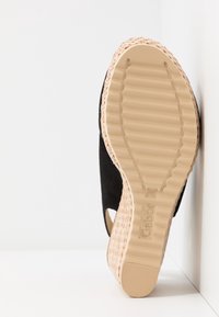 Semelle beige avec semelle striée et bordure tissée sur sandale compensée noire, positionnée verticalement contre un fond blanc d'angle.