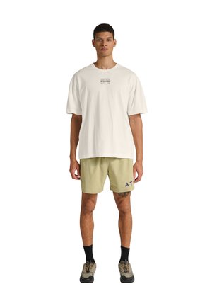 Junger Mann steht und trägt ein lockeres weißes T-Shirt mit Text, beige Shorts mit kleinem Muster, schwarze Socken und beige Turnschuhe.