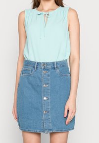 Top senza maniche verde menta con dettaglio a fiocco al collo, abbinato a una gonna di jeans blu con bottoni frontali e tasche.