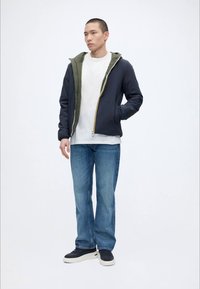 Homme portant une veste zippée noire et olive par-dessus une chemise blanche, un jean bleu et des baskets noires, debout, une main dans la poche.