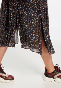 Ben i farverige chunky sneakers og en sort midi-længde blomsterkjole med sidelæg, stående på et lyst gulv.