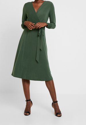 Femme portant une robe portefeuille à manches longues de couleur vert olive avec une ceinture à la taille, assortie à des sandales à talons hauts et brides à la cheville noires.